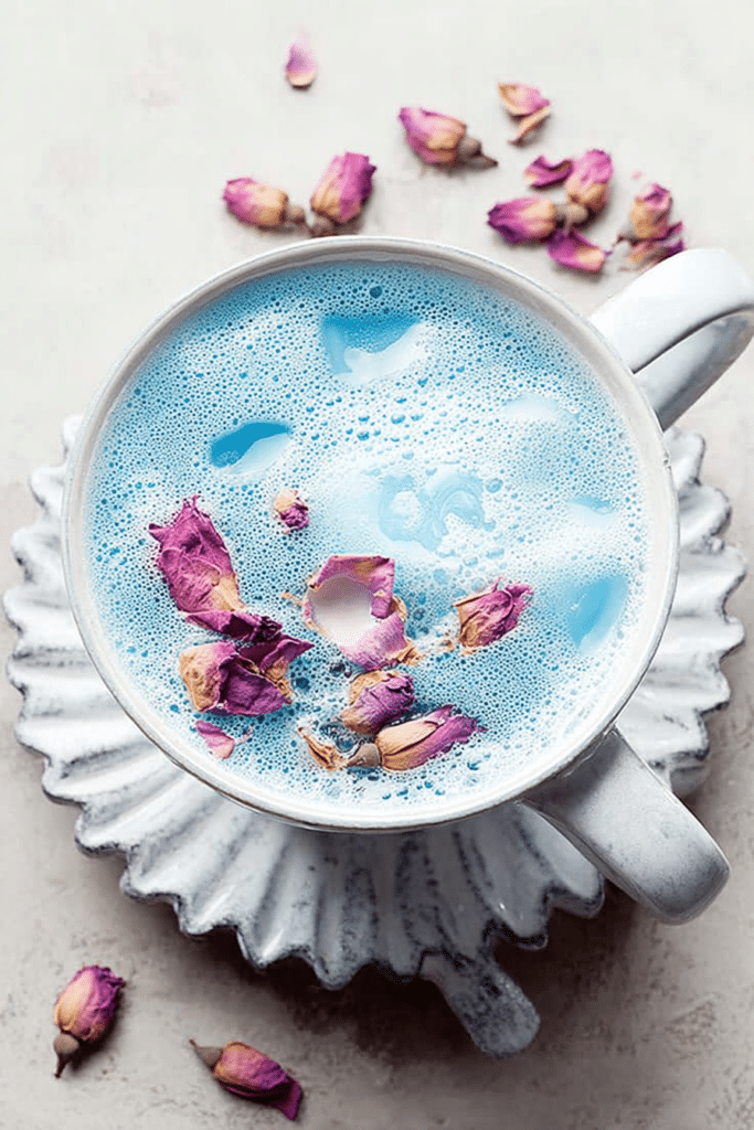 Close-up of a blue butterfly pea flower latte in a clear glass cup with rose petals on top, surrounded by blue butterfly pea flowers.