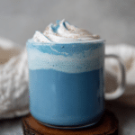 Blue latte with spirulina, topped with dairy-free whipped cream and cinnamon, served in a stylish mug on a rustic wooden table.