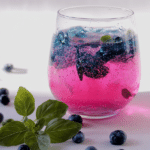 A glass of blueberry basil soda spritzer filled with ice, fresh blueberries, and basil leaves on a wooden table.