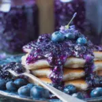 A jar of homemade blueberry lavender preserves surrounded by fresh blueberries and lavender flowers on a wooden surface.