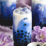 Glass of coconut butterfly pea flower boba tea with blue color and coconut cream layered, garnished with mint leaves.