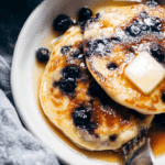 Fluffiest blueberry pancakes stacked high, topped with fresh blueberries and syrup on a wooden table.