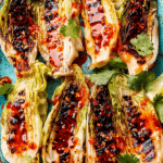 Grilled cabbage half topped with sweet chili sauce, lime wedges, and cilantro on a wooden table.