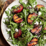 Grilled plum salad with goat cheese, arugula, and fresh herbs on a wooden table.