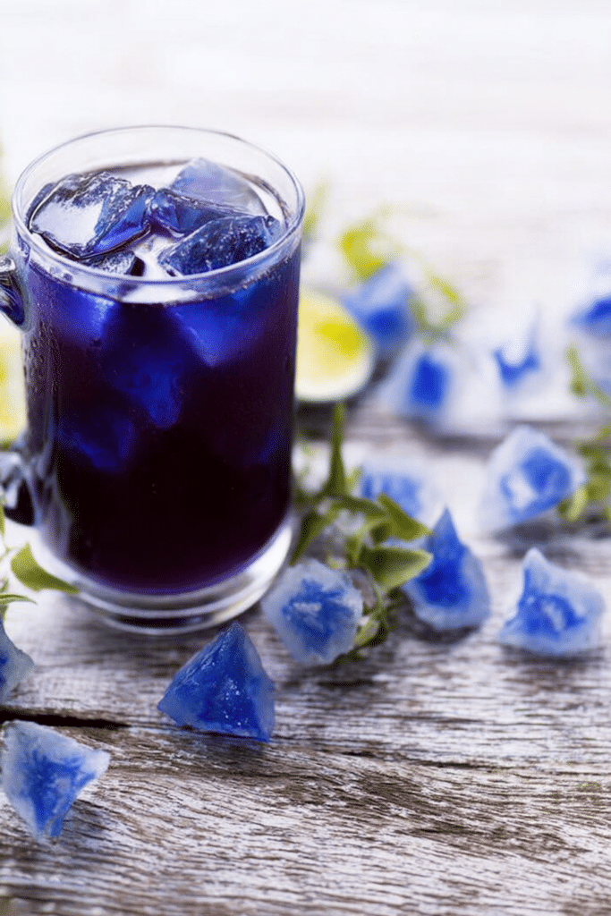 A refreshing glass of iced blue tea garnished with a lemon slice, featuring ingredients like blue tea flowers, chamomile, and ice.