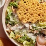 Micro chop caesar salad with grilled chicken, croutons, and a giant parmesan crisp in a white bowl.