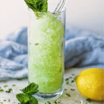 Mint lemonade slushie in a glass with mint leaves and lemon slices, served with ice on a wooden table.