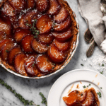 Slice of plum & thyme tarte tatin with fresh thyme and plums on a wooden table.