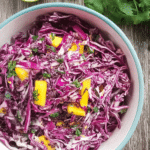 Bowl of colorful red cabbage and mango slaw with cilantro on a wooden table, surrounded by whole mangoes and red cabbage.