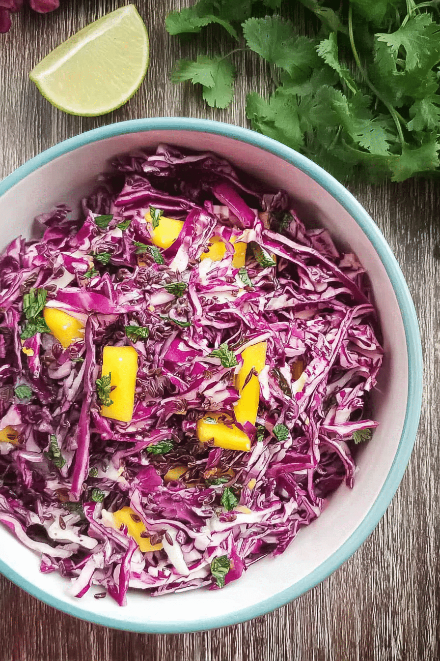 Bowl of colorful red cabbage and mango slaw with cilantro on a wooden table, surrounded by whole mangoes and red cabbage.