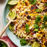 Colorful street corn slaw in a bowl with crumbled cotija cheese and lime wedges, garnished with cilantro.